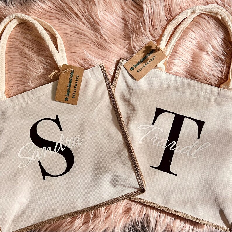 K&ouml;nnte beinhalten: Zwei beigefarbene Tragetaschen mit cremefarbenen Henkeln und goldfarbenem Besatz. Eine Tasche hat ein gro&szlig;es schwarzes "S" mit dem Namen "Sandra" in wei&szlig;er Schrift, die andere ein schwarzes "T" mit dem Namen "Traudl". Jede Tasche hat ein braunes Etikett.
