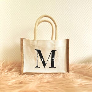 May include: A white canvas tote bag with brown jute trim. The bag has a single black letter "M" printed on the front.