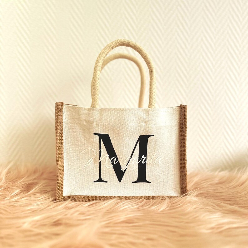 May include: A white canvas tote bag with brown jute trim. The bag has a single black letter "M" printed on the front.