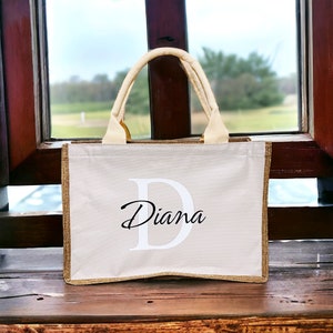K&ouml;nnte beinhalten: Beige Tragetasche mit goldenem Besatz und cremefarbenen Griffen. Die Tasche hat einen gro&szlig;en wei&szlig;en Buchstaben "D" und den Namen "Diana" in Schreibschrift. Die Tasche steht auf einer Holzoberfl&auml;che.