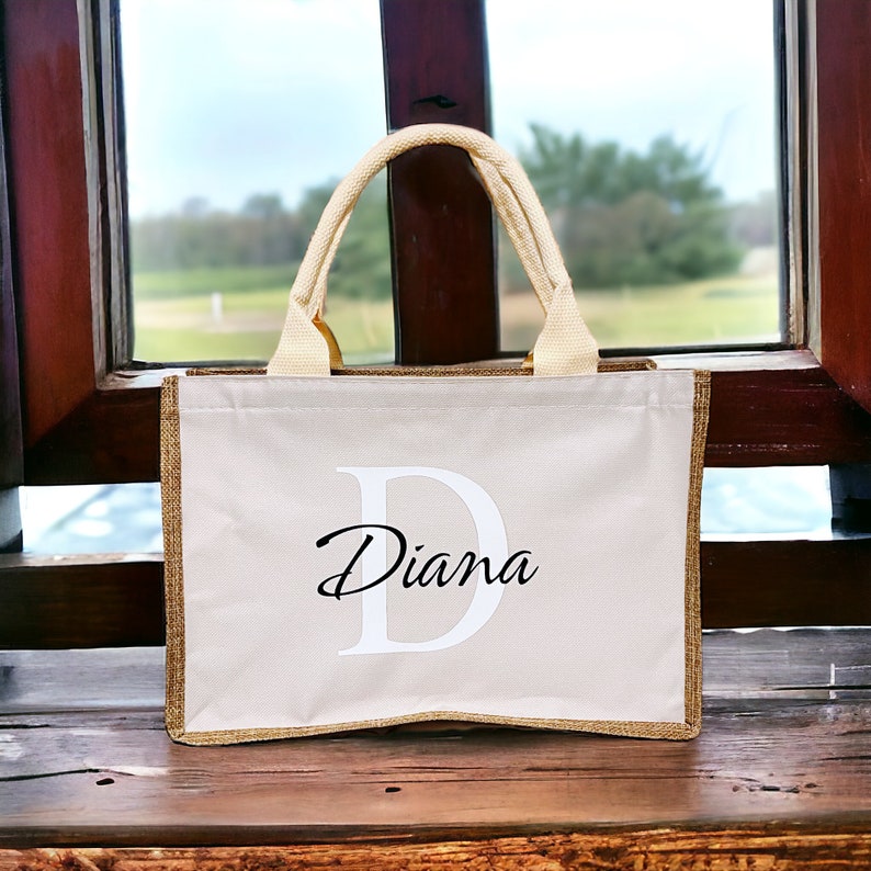 K&ouml;nnte beinhalten: Beige Tragetasche mit goldenem Besatz und cremefarbenen Griffen. Die Tasche hat einen gro&szlig;en wei&szlig;en Buchstaben "D" und den Namen "Diana" in Schreibschrift. Die Tasche steht auf einer Holzoberfl&auml;che.