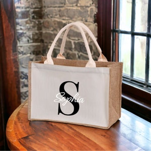 K&ouml;nnte beinhalten: Rechteckige Jute-Tragetasche mit wei&szlig;er Frontplatte. Die Frontplatte zeigt einen gro&szlig;en schwarzen Buchstaben "S" und den Namen "Sophia" in Schreibschrift. Die Tasche hat beige Griffe und einen beigen K&ouml;rper.