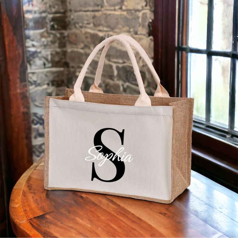 K&ouml;nnte beinhalten: Rechteckige Jute-Tragetasche mit wei&szlig;er Frontplatte. Die Frontplatte zeigt einen gro&szlig;en schwarzen Buchstaben "S" und den Namen "Sophia" in Schreibschrift. Die Tasche hat beige Griffe und einen beigen K&ouml;rper.
