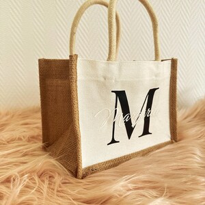 May include: A small, white canvas tote bag with brown burlap sides. The bag has a single, white rope handle and features a black letter "M" printed on the front.