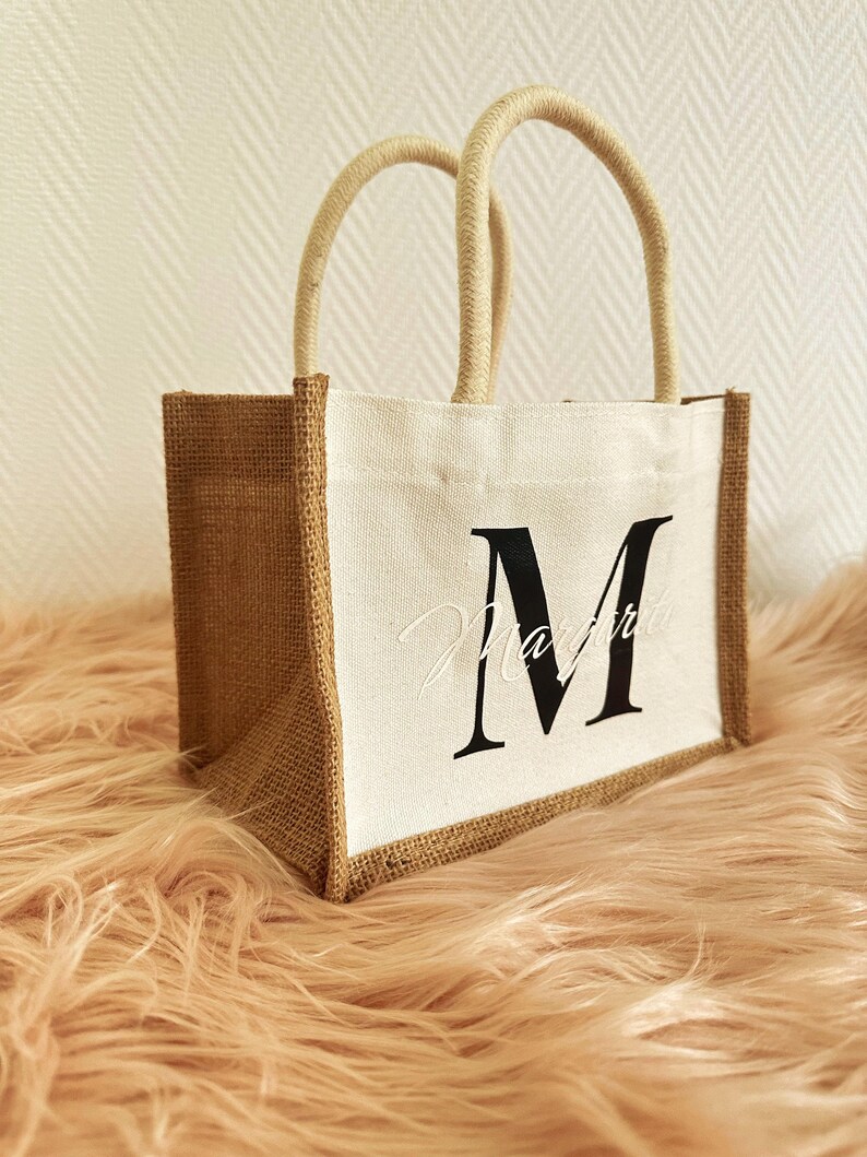 May include: A small, white canvas tote bag with brown burlap sides. The bag has a single, white rope handle and features a black letter "M" printed on the front.