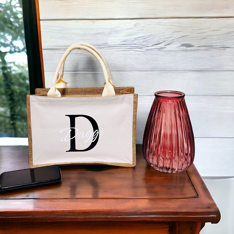 K&ouml;nnte beinhalten: Eine beigefarbene Tragetasche mit einer wei&szlig;en Frontplatte, die den gro&szlig;en schwarzen Buchstaben "D" und den Namen "Daggi" in Schreibschrift zeigt. Die Tasche hat einen K&ouml;rper aus Jute, cremefarbene Griffe und eine passende Einfassung. Eine rote Glasvase und ein Smartphone befinden sich ebenfalls auf der Holzoberfl&auml;che.