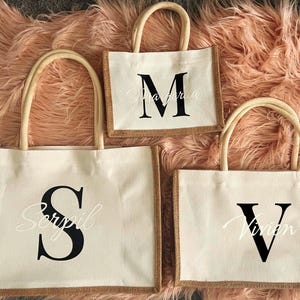 May include: Three natural fibre tote bags with brown trim. The largest bag has a black letter "S" with the name "Serpil" in white script. The medium bag has a black letter "M" with the name "Margarita" in white script. The smallest bag has a black letter "V" with the name "Vivian" in white script.