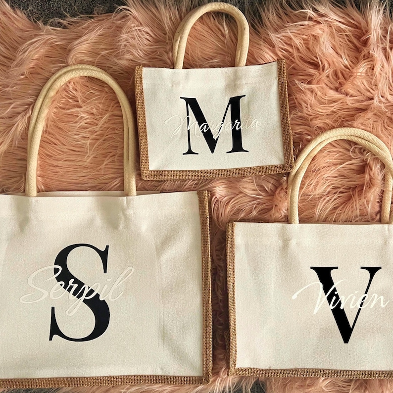 May include: Three natural fibre tote bags with brown trim. The largest bag has a black letter "S" with the name "Serpil" in white script. The medium bag has a black letter "M" with the name "Margarita" in white script. The smallest bag has a black letter "V" with the name "Vivian" in white script.