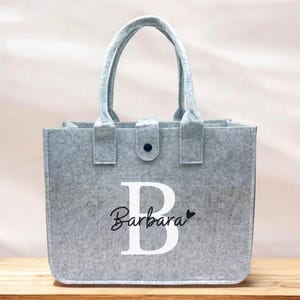 Könnte beinhalten: Eine graue Filz-Tragetasche mit Griff und Knopfverschluss. Die Tasche trägt den Namen "Barbara" in einer Schreibschrift mit einem großen weißen Buchstaben "B". Neben dem Namen befindet sich ein kleines schwarzes Herz.