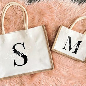 May include: Two personalised tote bags with burlap trim. The larger bag has a white background with a black letter "S" and the text "Serpil" underneath. The smaller bag has a white background with a black letter "M".