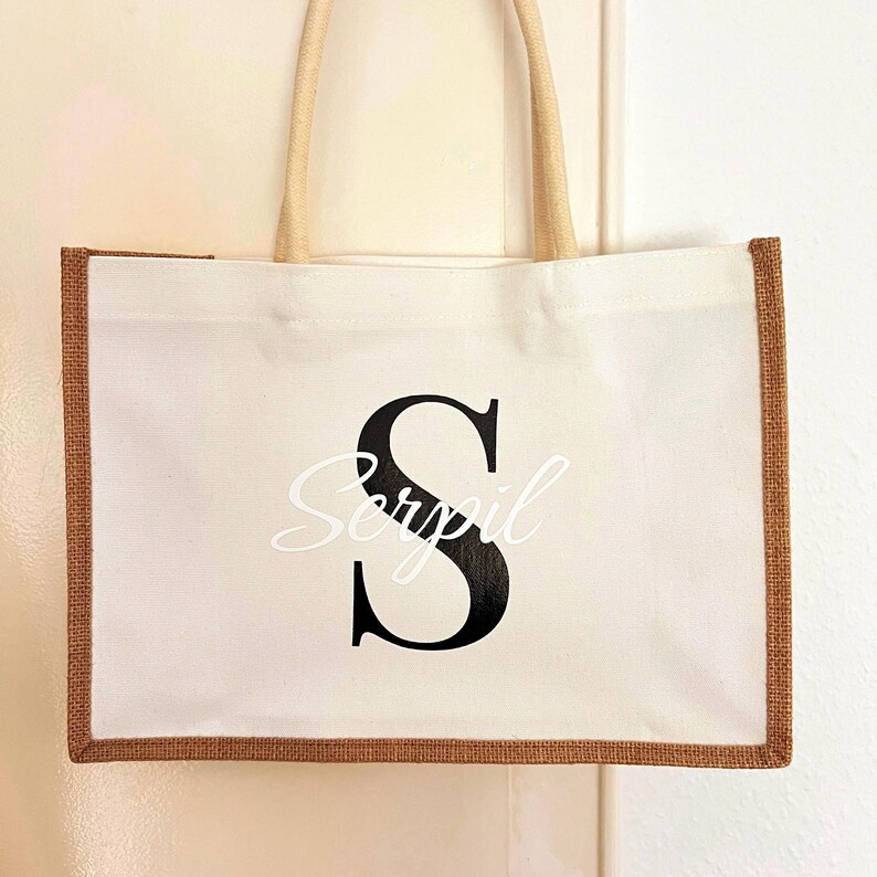 May include: A white canvas tote bag with brown jute trim. The bag has a black letter "S" and the name "Serypil" printed on the front.