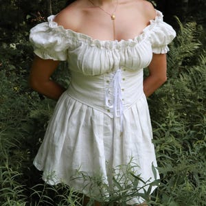 May include: A white off-the-shoulder linen dress with a fitted bodice and a lace-up corset detail. The dress has a gathered skirt and short puff sleeves.