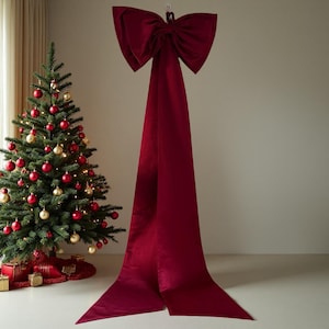 Könnte beinhalten: Eine große, tiefrote dekorative Schleife mit langen, fließenden Bändern hängt vor einer neutralen Wand. Links steht ein Weihnachtsbaum, geschmückt mit roten und goldenen Kugeln und Lichtern, was eine festliche Szene schafft.