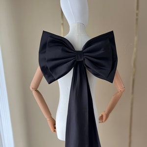 Op de afbeelding: Een grote zwarte satijnen strik met een lang, hangend lint. De strik is om een wit mannequin gebonden.