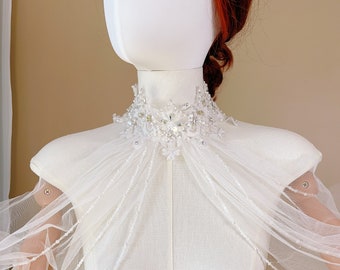 Puede incluir: Un chal de novia de tul blanco con un cuello de cuentas y una franja de cuentas. El chal está hecho de tul delicado y presenta un hermoso diseño de cuentas.