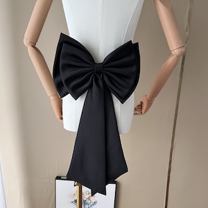 Op de afbeelding: Een grote, zwarte satijnen strik met lange, vloeiende linten. De strik is bevestigd aan een witte mannequin torso, die de vorm en het ontwerp benadrukt. De stof van de strik heeft een subtiele glans en de linten lopen naar beneden.