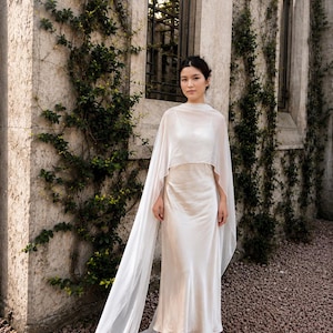 May include: A floor-length, cream-coloured satin slip dress featuring a scoop neckline and thin spaghetti straps, paired with a sheer, lightweight, floor-length cape that drapes over the shoulders and extends into a long train behind the garment.