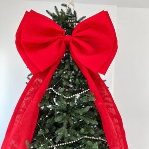 Puede incluir: Un gran lazo de tela de color rojo vibrante que sirve como adorno para la punta del árbol de Navidad. El lazo presenta dos cintas anchas y fluidas que caen por los lados del pino verde, decorado con delicadas hileras de cuentas blancas.