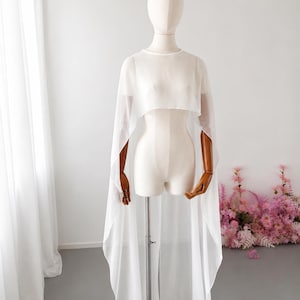 May include: A sheer white cape-style garment with a high neckline and flowing, floor-length design. The cape is displayed on a mannequin with wooden arms, showcasing its delicate fabric and elegant drape. The background features a white wall and pink flowers.