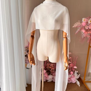 May include: A sheer white chiffon capelet draped over a mannequin. The garment features a high neckline and long, flowing panels that extend down from the shoulders, creating a layered effect with translucent fabric that hangs loosely.