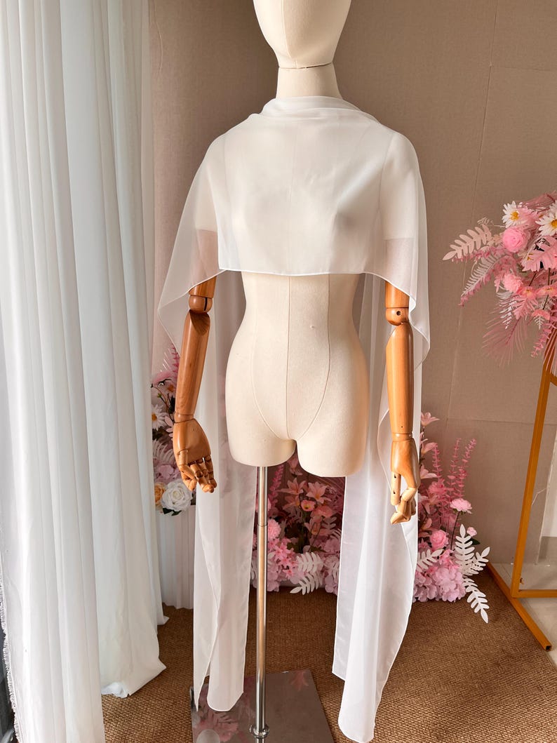 May include: A sheer white chiffon capelet draped over a mannequin. The garment features a high neckline and long, flowing panels that extend down from the shoulders, creating a layered effect with translucent fabric that hangs loosely.