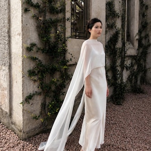 May include: A floor-length white slip dress featuring a sheer, lightweight overlay that drapes over the shoulders and extends into long, flowing panels. The garment has a high neckline, thin spaghetti straps underneath, and a smooth, satin-like finish.