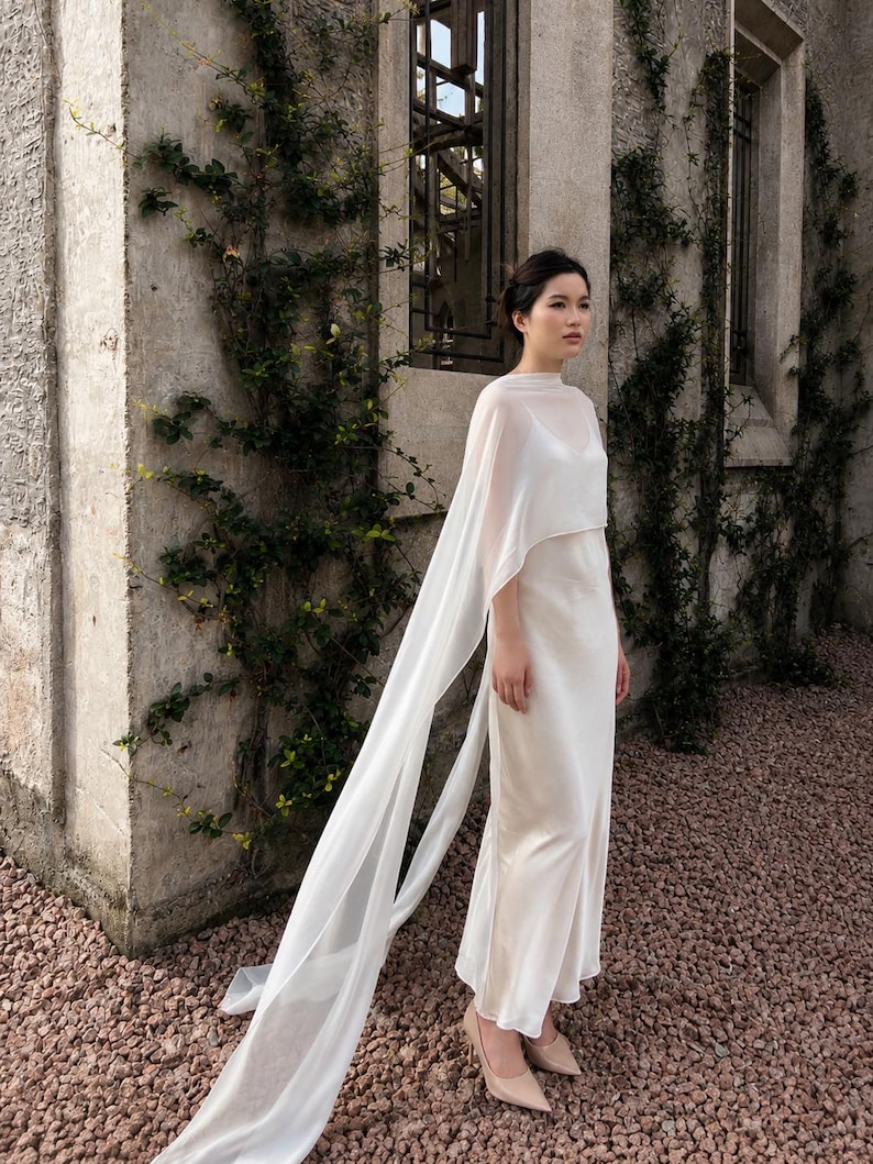 May include: A floor-length white slip dress featuring a sheer, lightweight overlay that drapes over the shoulders and extends into long, flowing panels. The garment has a high neckline, thin spaghetti straps underneath, and a smooth, satin-like finish.