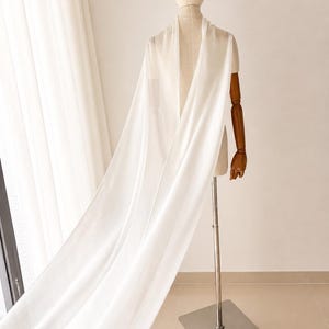 May include: A white, flowing fabric draped over a mannequin. The fabric falls in soft folds, creating a sense of movement. The mannequin has wooden arms and a neutral-toned head. The background is a light, neutral colour.