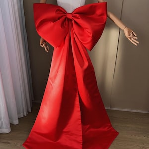 May include: A vibrant red satin bow and train, displayed on a mannequin. The bow is large and voluminous, with a flowing train extending downwards. The fabric has a glossy sheen, and the overall effect is dramatic and eye-catching. The bow is a statement piece.