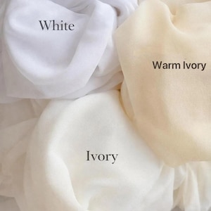 May include: Three swatches of sheer fabric in different shades labelled White, Warm Ivory, and Ivory. The text Available Colours is printed at the bottom. The fabric has a soft, draped texture with visible folds and light-filtering properties.