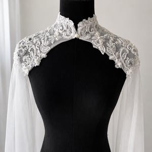 May include: A white bridal capelet featuring a high collar and intricate floral embroidery with silver beads, sequins, and small pearls. The garment is crafted from sheer tulle fabric that drapes down from the shoulders, secured by a single pearl button.