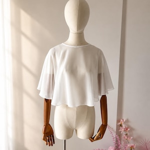 Peut inclure: Un chemisier blanc semi-transparent à manches courtes, doté d'une encolure ronde et d'une coupe courte et évasée. Le vêtement est composé d'un tissu léger et fluide qui tombe souplement depuis les épaules, créant un aspect doux et superposé.