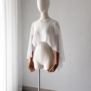 May include: A white, semi-sheer, cropped capelet featuring a wide, rounded neckline and flowing, open-front panels that drape over the shoulders and upper torso. The lightweight fabric creates a soft, layered effect with a high-low hemline.