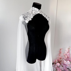 May include: A white sheer tulle cape featuring a high collar and intricate silver floral embroidery with beadwork embellishments across the shoulders and neckline. The cape drapes down from the shoulders in long, flowing panels of lightweight fabric.