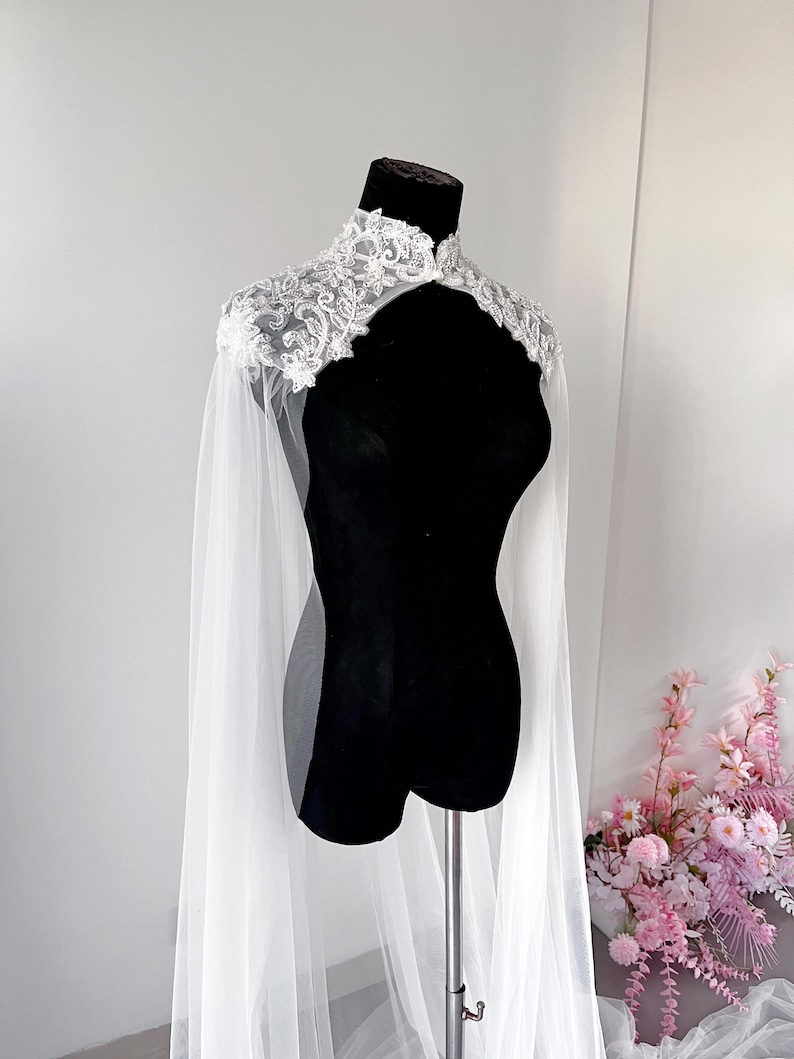May include: A white sheer tulle cape featuring a high collar and intricate silver floral embroidery with beadwork embellishments across the shoulders and neckline. The cape drapes down from the shoulders in long, flowing panels of lightweight fabric.