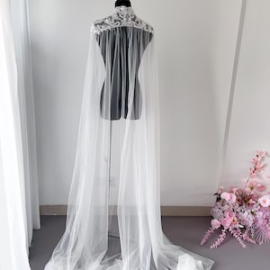 May include: A floor-length white tulle cape featuring a high collar adorned with intricate white floral lace embroidery and beadwork. The sheer, lightweight fabric drapes from the shoulders, creating a long, flowing silhouette that pools on the floor.