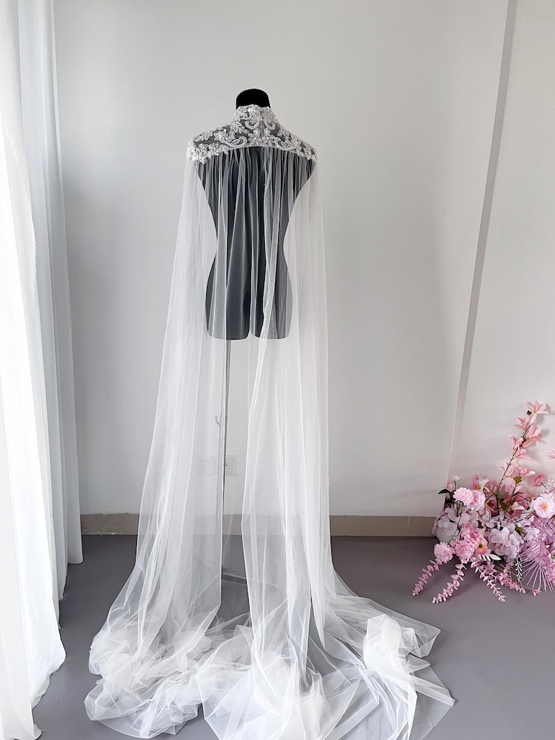 May include: A floor-length white tulle cape featuring a high collar adorned with intricate white floral lace embroidery and beadwork. The sheer, lightweight fabric drapes from the shoulders, creating a long, flowing silhouette that pools on the floor.
