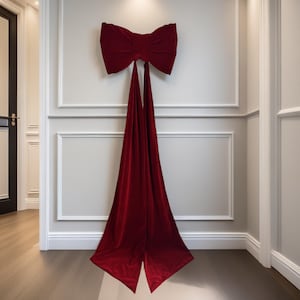 May include: A large, plush, burgundy velvet bow is mounted on a white wall with decorative moulding. The bow's long, flowing tails extend downwards, creating a dramatic and elegant decorative accent. The bow is a striking focal point in the room.