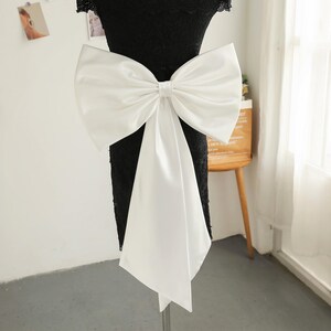 Op de afbeelding: Een witte satijnen strik met lange linten is bevestigd aan een zwarte kanten jurk. De strik is een opvallend kenmerk, met een klassiek ontwerp. De jurk wordt op een mannequin gepresenteerd, wat de elegante stijl van de strik benadrukt.