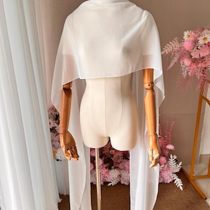 May include: A sheer white chiffon capelet draped over a mannequin. The garment features a high neckline and long, flowing panels that extend down from the shoulders, creating a lightweight, semi-transparent layer with a smooth, matte finish.