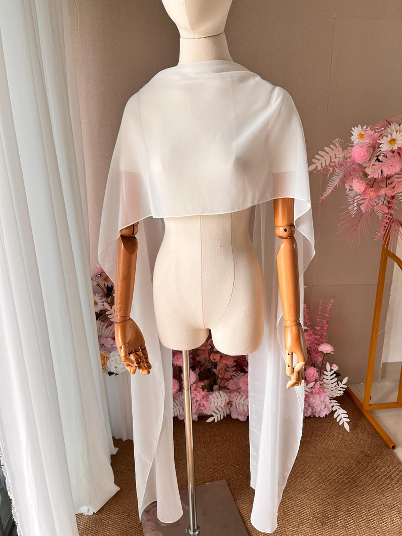 May include: A sheer white chiffon capelet draped over a mannequin. The garment features a high neckline and long, flowing panels that extend down from the shoulders, creating a lightweight, semi-transparent layer with a smooth, matte finish.