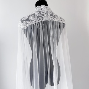 May include: A white sheer tulle cape featuring an intricate high-neck collar adorned with silver-toned floral embroidery, sequins, and beads. The cape drapes from the shoulders with a flowing, translucent fabric design that extends downward.