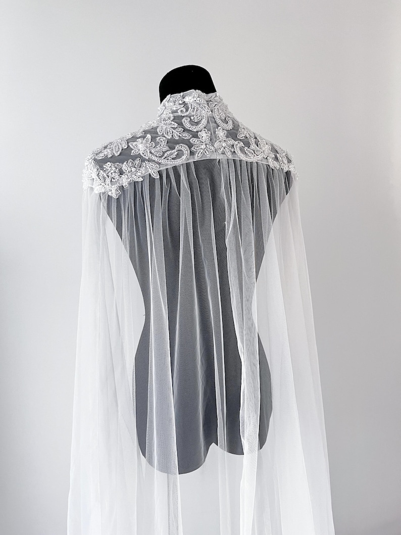 May include: A white sheer tulle cape featuring an intricate high-neck collar adorned with silver-toned floral embroidery, sequins, and beads. The cape drapes from the shoulders with a flowing, translucent fabric design that extends downward.