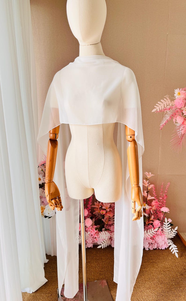 May include: A sheer white chiffon capelet draped over a mannequin. The garment features a high neckline and long, flowing panels that extend from the shoulders down past the waist, creating a lightweight, semi-transparent layered effect.