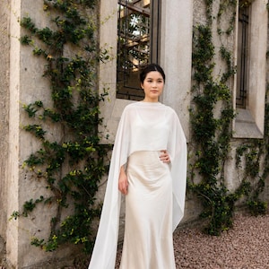 May include: A floor-length cream-coloured satin slip dress paired with a sheer white chiffon capelet. The capelet features long, flowing panels that drape down to the floor, while the dress has a simple scoop neckline and a smooth, lustrous finish.