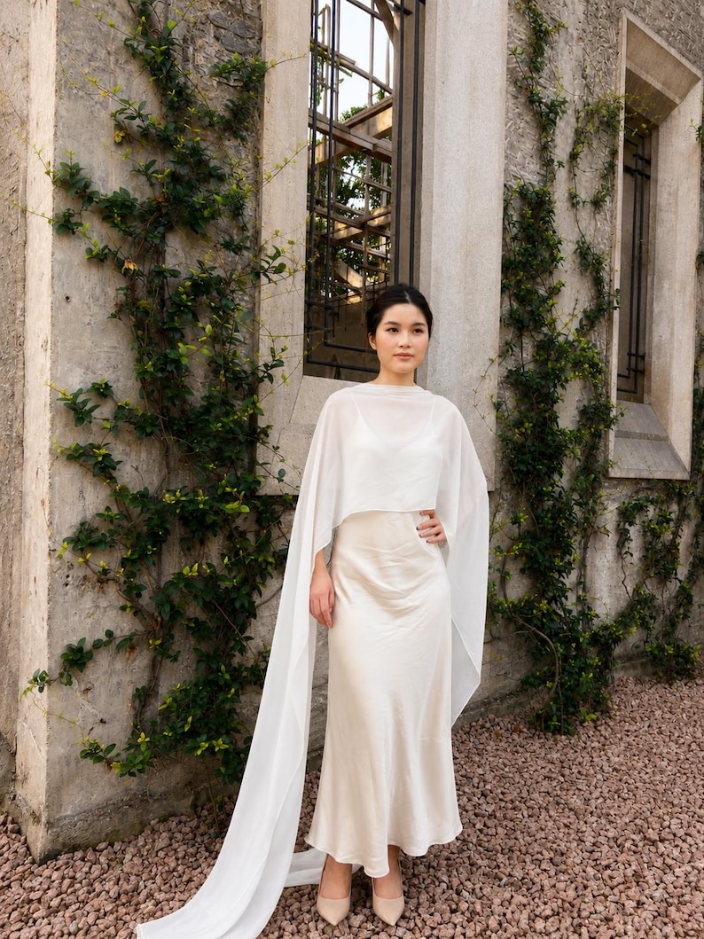 May include: A floor-length cream-coloured satin slip dress paired with a sheer white chiffon capelet. The capelet features long, flowing panels that drape down to the floor, while the dress has a simple scoop neckline and a smooth, lustrous finish.