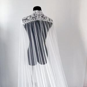 May include: A floor-length white bridal cape featuring a high collar adorned with intricate silver embroidery, sequins, and beadwork. The cape is constructed from sheer, lightweight tulle fabric that drapes from the shoulders to the floor.