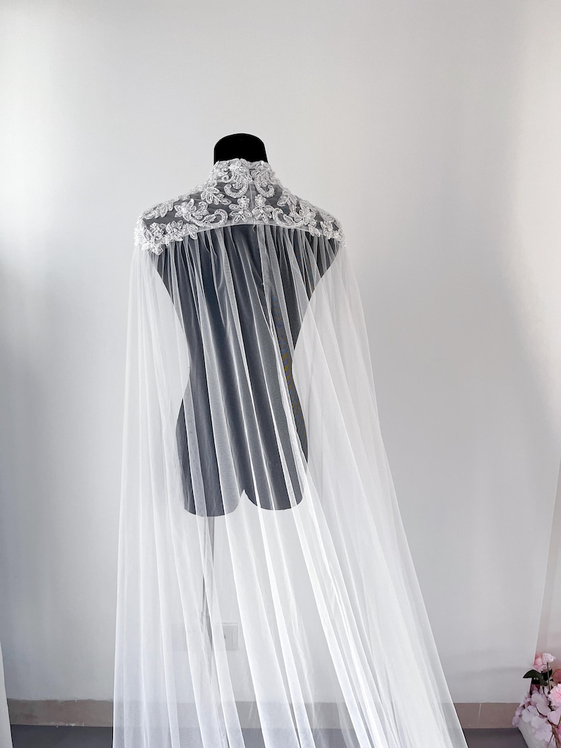 May include: A floor-length white bridal cape featuring a high collar adorned with intricate silver embroidery, sequins, and beadwork. The cape is constructed from sheer, lightweight tulle fabric that drapes from the shoulders to the floor.