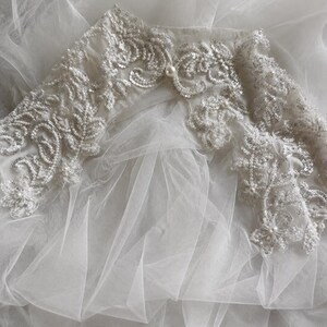 May include: A white tulle garment featuring an intricate embroidered collar adorned with silver sequins, clear beads, and small white pearls. The sheer, layered fabric creates a soft, voluminous texture beneath the detailed floral-inspired embellishments.