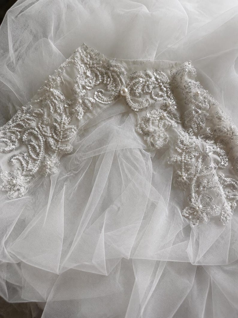 May include: A white tulle garment featuring an intricate embroidered collar adorned with silver sequins, clear beads, and small white pearls. The sheer, layered fabric creates a soft, voluminous texture beneath the detailed floral-inspired embellishments.